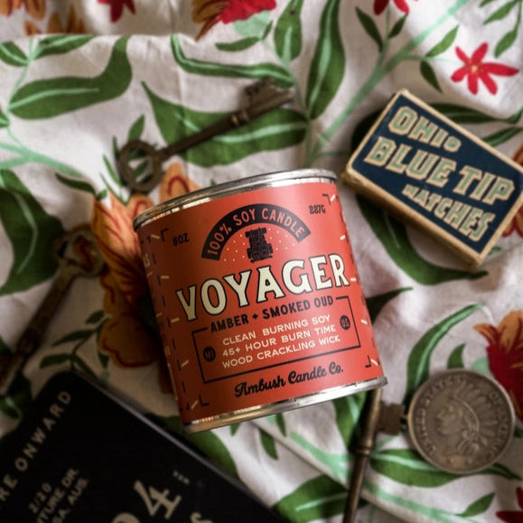Voyager candle tin on a floral fabric background with a matchbox and coin.