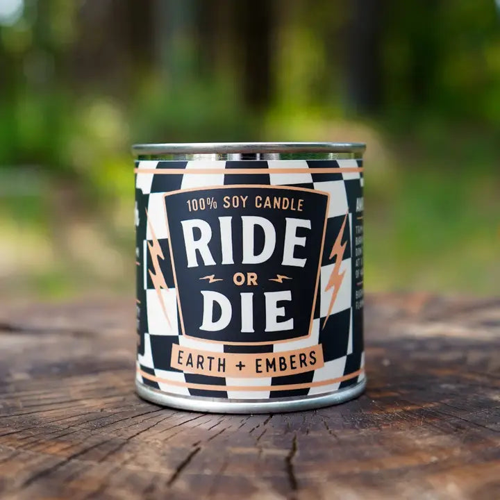 Candle in a can labeled 'Ride or Die' on a wooden surface with a blurred natural background